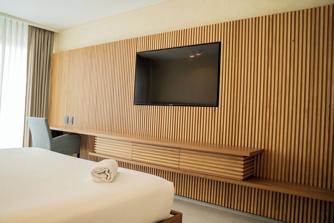 A modern hotel room with a large bed, a flat-screen TV mounted on a wooden slat wall, and a seating area with a bench.