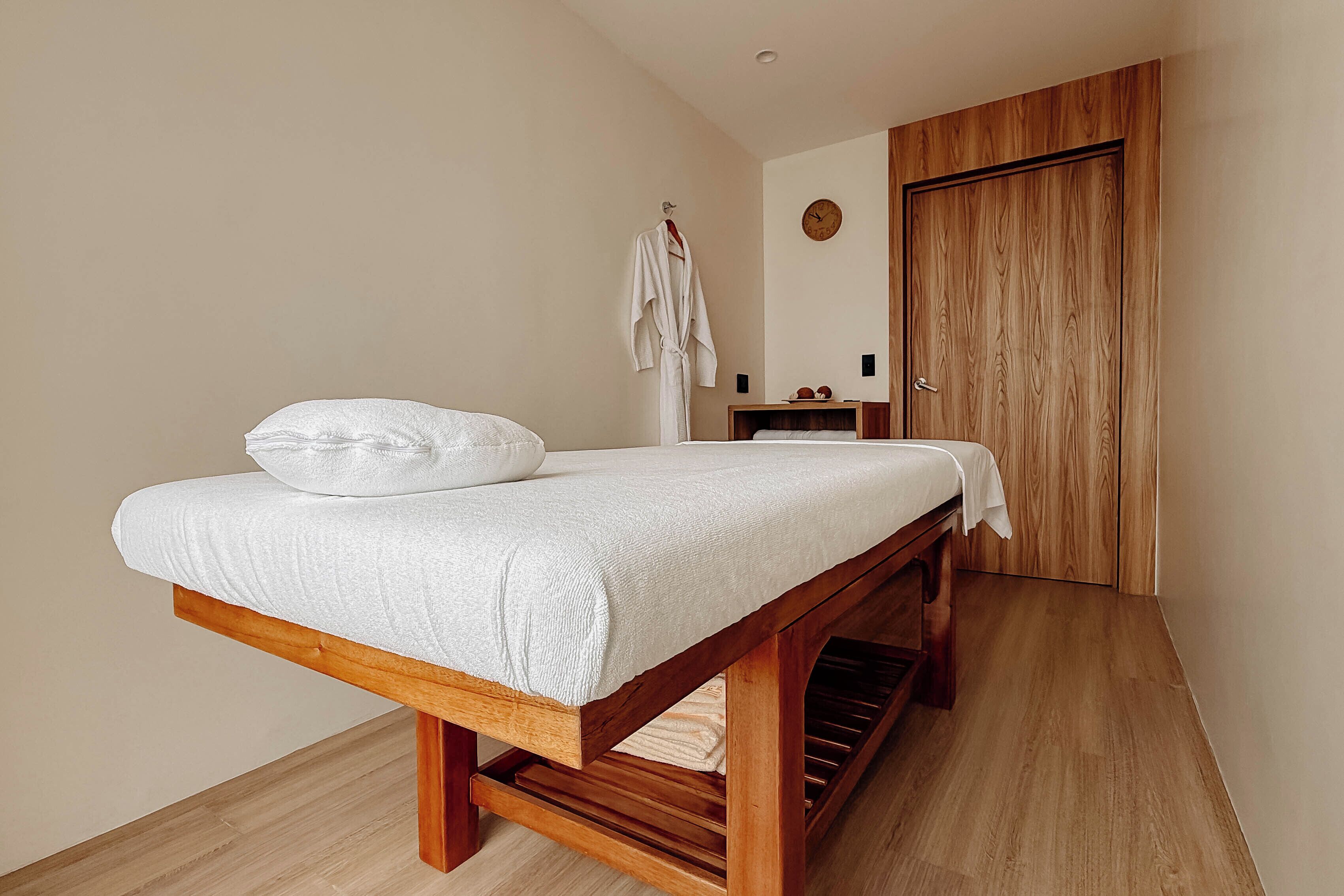 A room with a wooden bed, a white robe hanging on a hook, a wooden wardrobe, and a small bedside table with a clock.