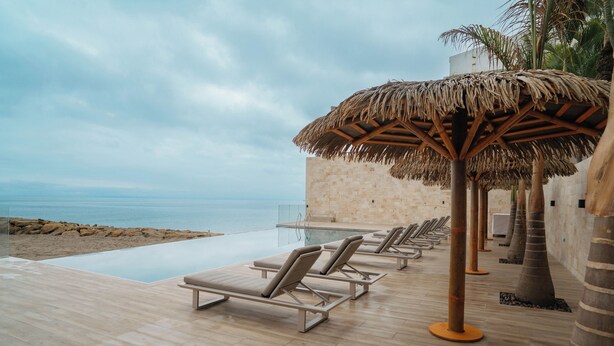 A poolside area with lounge chairs and a thatched umbrella, overlooking a beach and the sea.