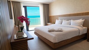 A modern hotel room with a large bed, a wooden nightstand, a bedside table with a vase of flowers, and a view of the ocean through a large window.