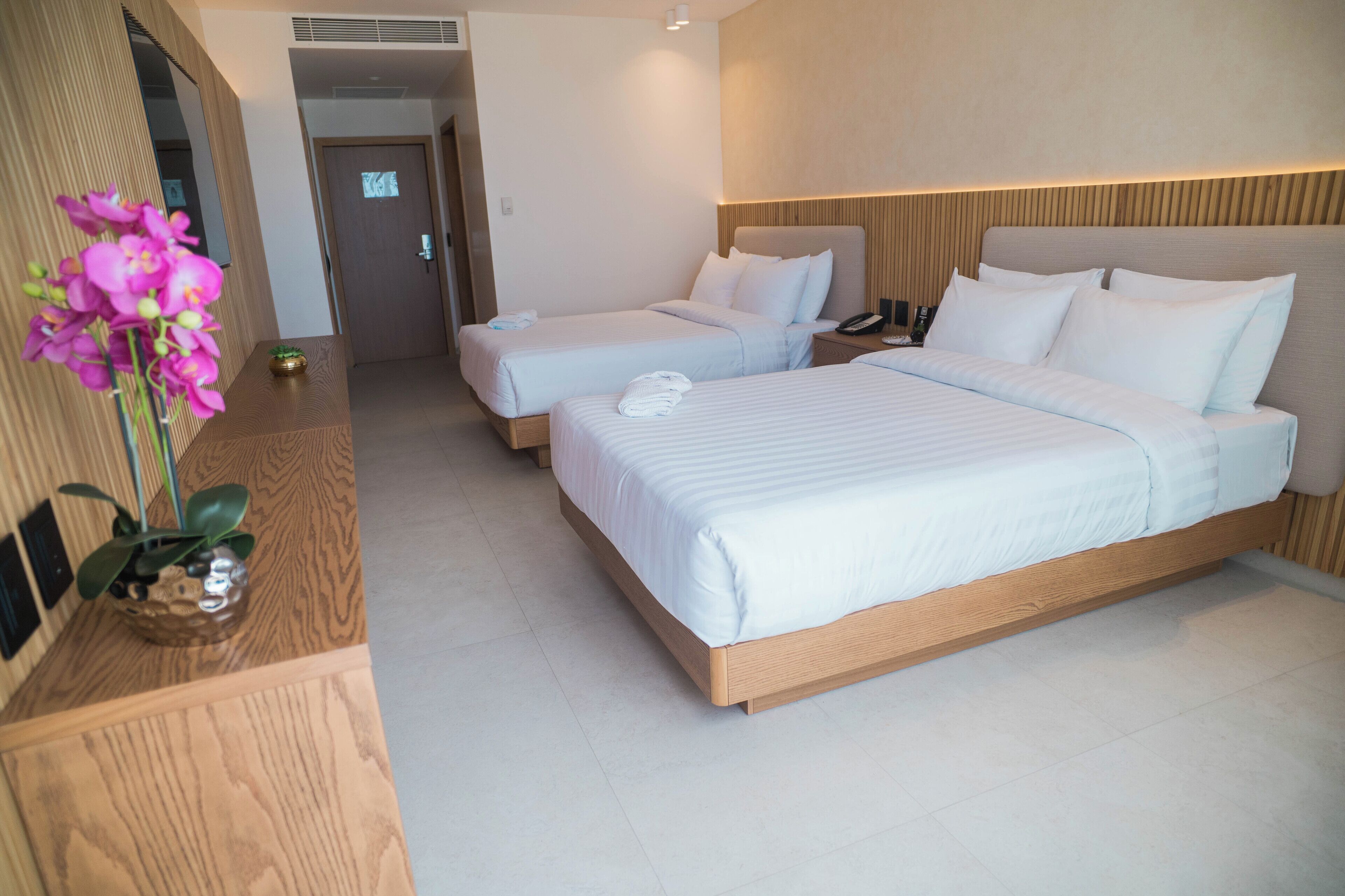 A hotel room with two beds, a wooden headboard, a bedside table with a lamp, and a potted orchid.