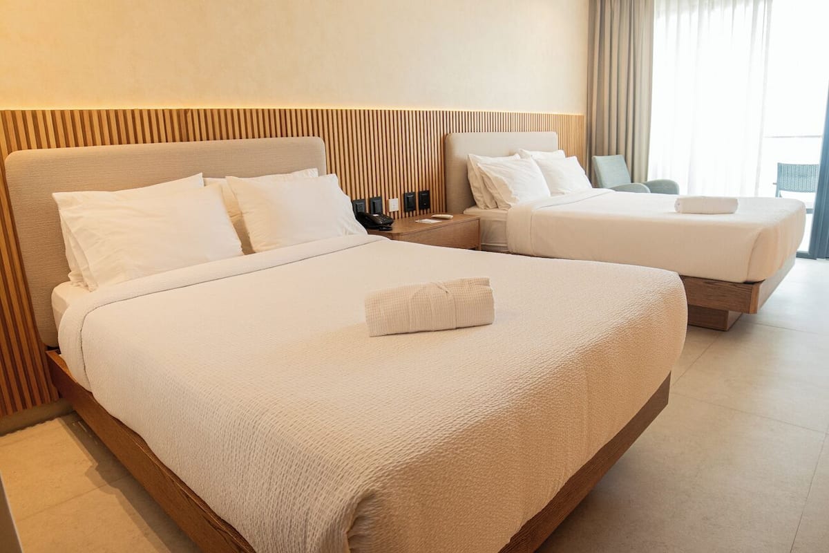 A hotel room with two beds, each with white linens and a wooden headboard. There is a bedside table with a lamp and a phone on one side.