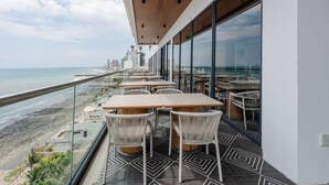 A balcony with tables and chairs overlooking a beach and cityscape.