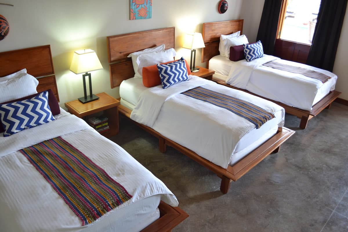 Une chambre d’hôtel avec trois lits simples, chacun doté d’une tête de lit en bois, de draps blancs et de coussins décoratifs aux couleurs vives. On y trouve des tables de chevet avec des lampes, et le sol est carrelé.