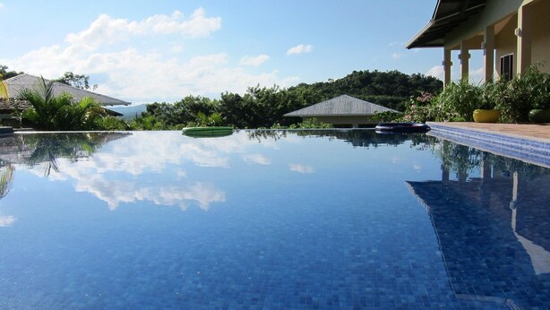 A clear swimming pool with a tiled edge, surrounded by lush greenery and a building with a tiled roof. - Soma Surf Resort (Tola)