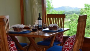 A wooden dining table with blue cushioned chairs, a bottle of wine, and a scenic view of green hills. - Soma Surf Resort (Tola)