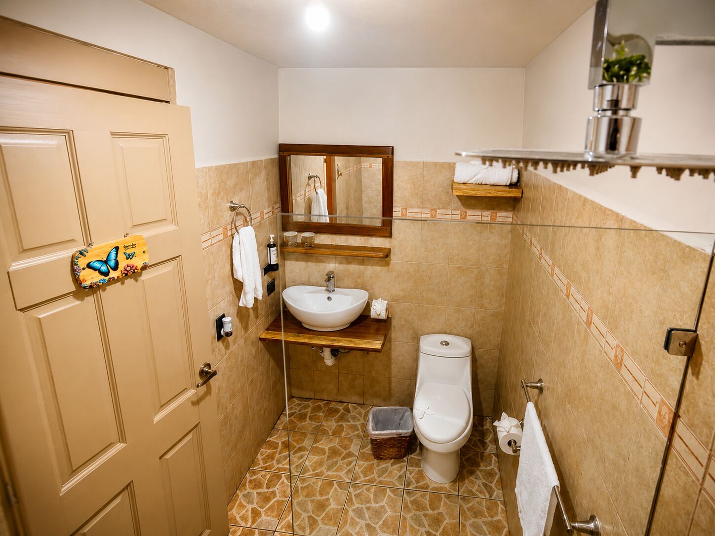 A bathroom with a sink, toilet, and mirror. There is a towel rack, a small shelf with toiletries, and a decorative butterfly sign on the door.