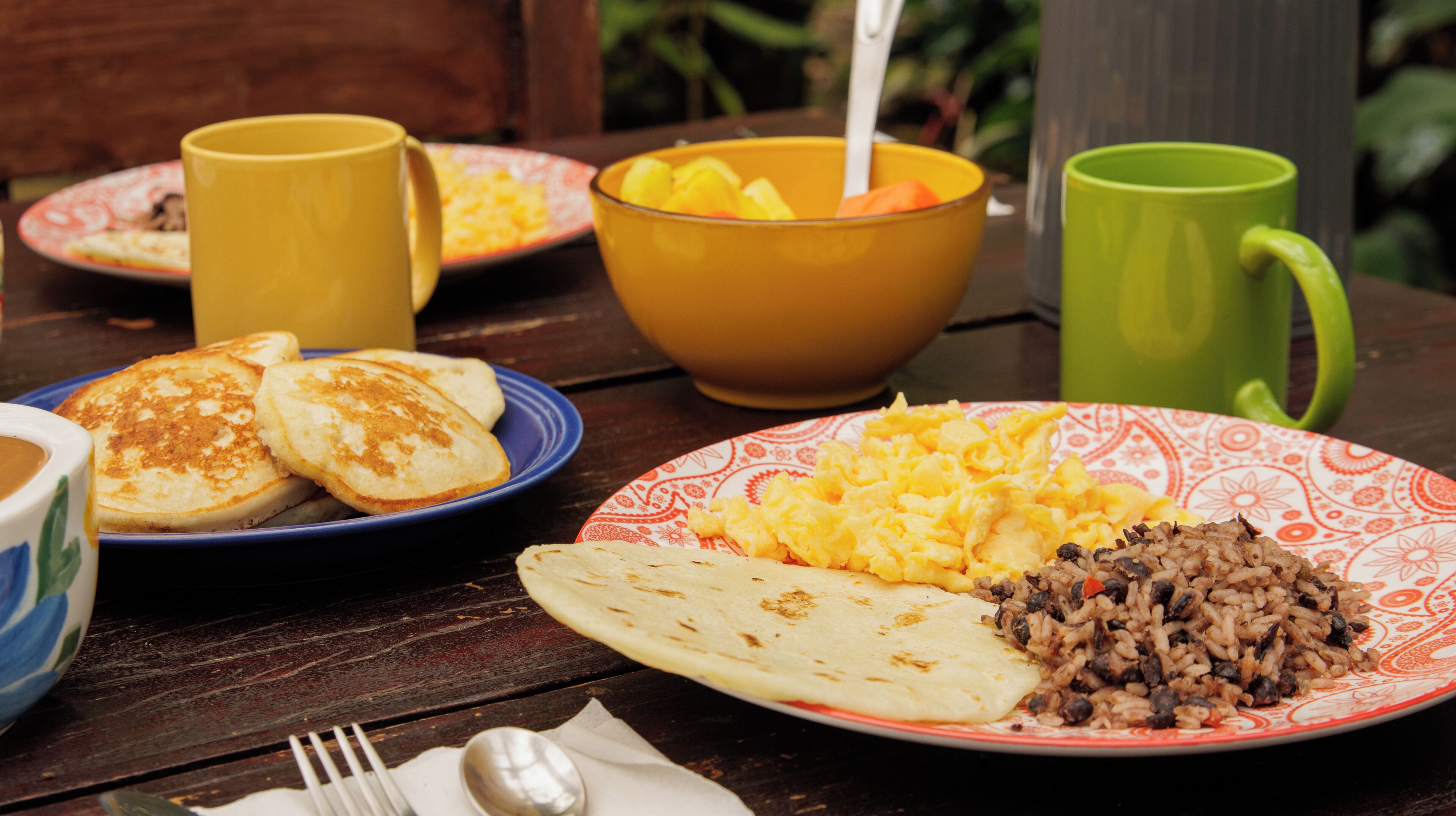 A table with breakfast plates, including pancakes, scrambled eggs, rice, and a tortilla. There are also two mugs and a bowl with fruit.