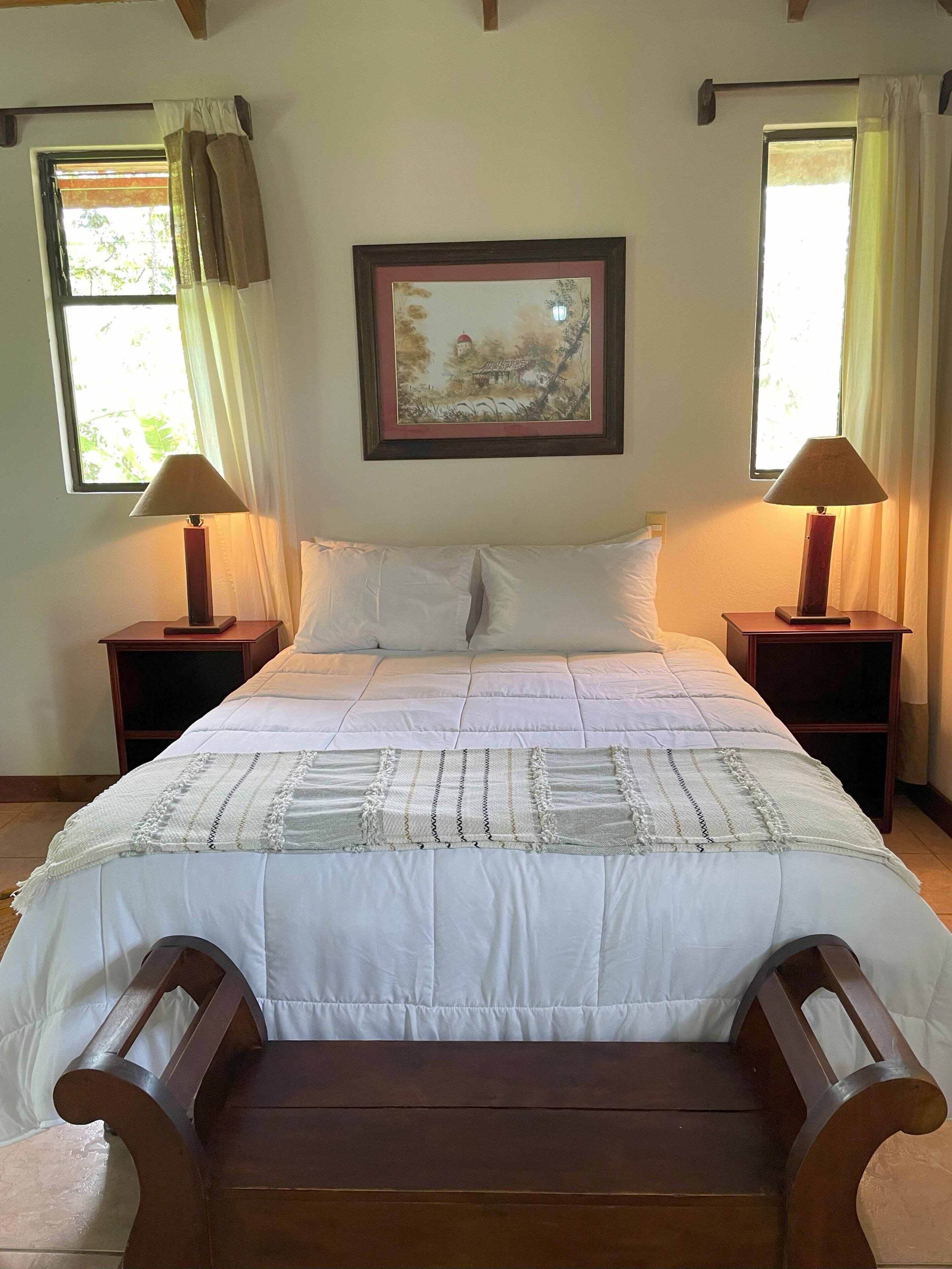 A neatly made bed with a white comforter and a decorative throw blanket. There are two wooden nightstands with lamps and a framed picture above the bed.