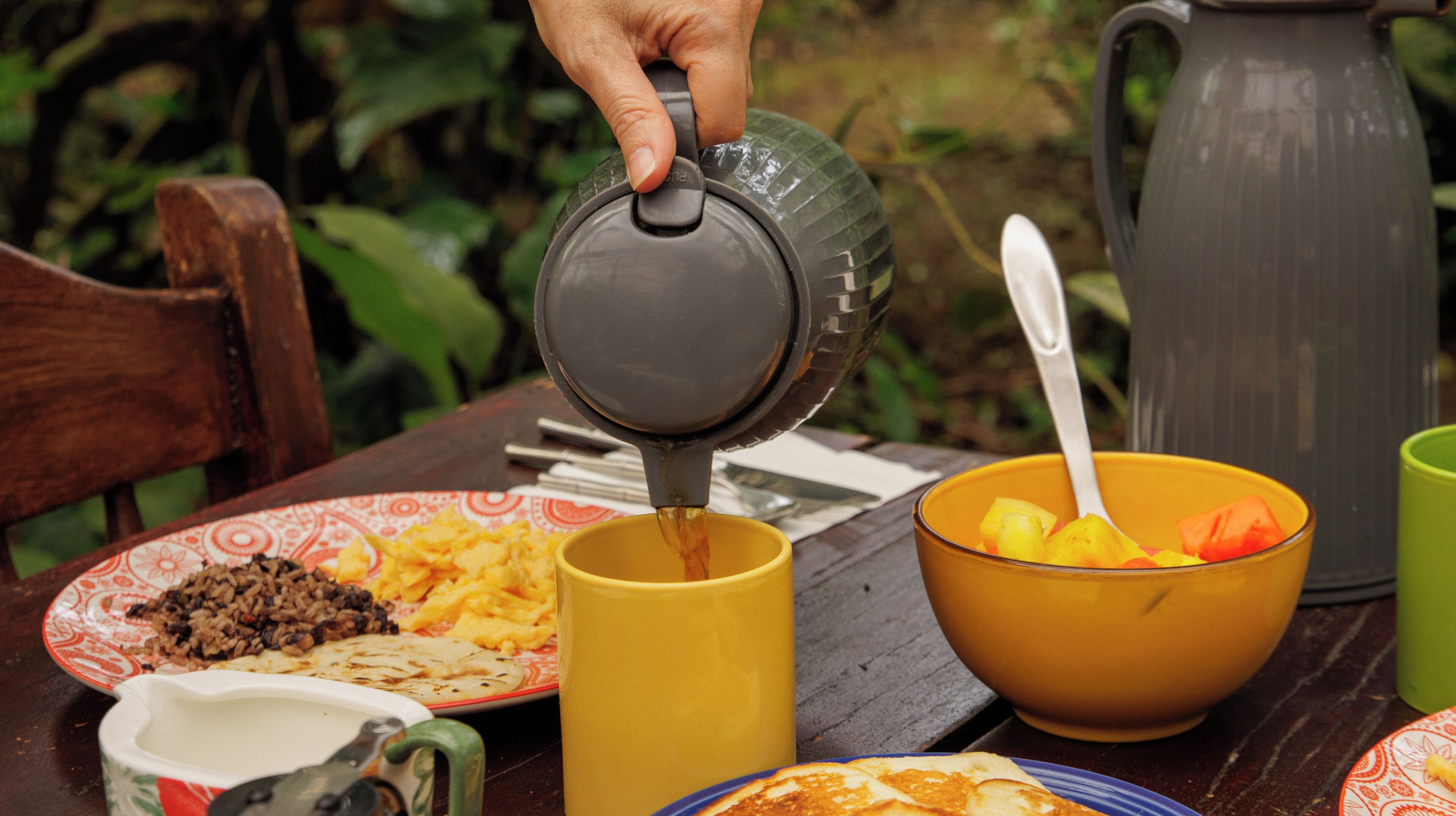 Eine Hand gießt ein Getränk in eine gelbe Tasse, eine Schüssel mit Obst und verschiedene Gerichte stehen auf einem Tisch.