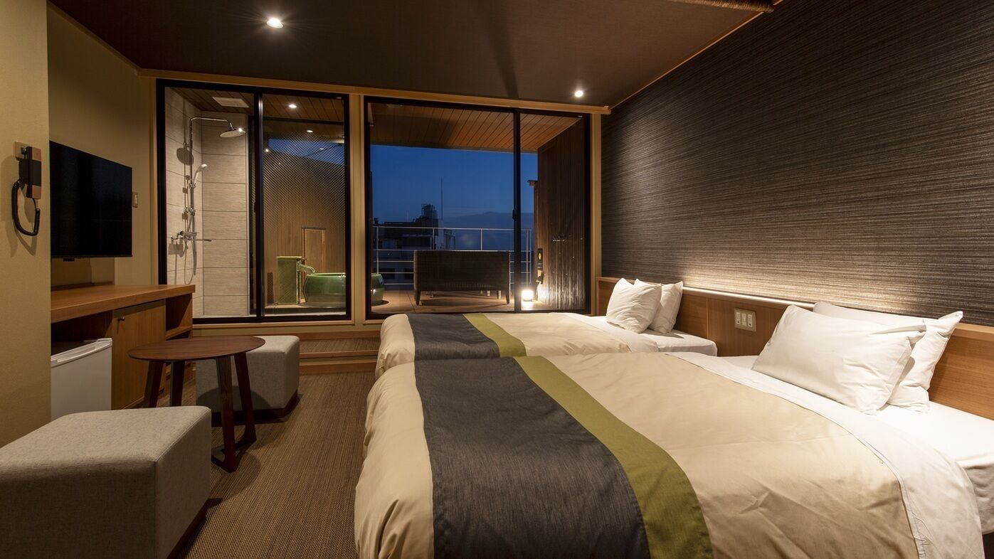 A hotel room with two beds, a desk, a chair, a TV, and a sliding glass door leading to a balcony.