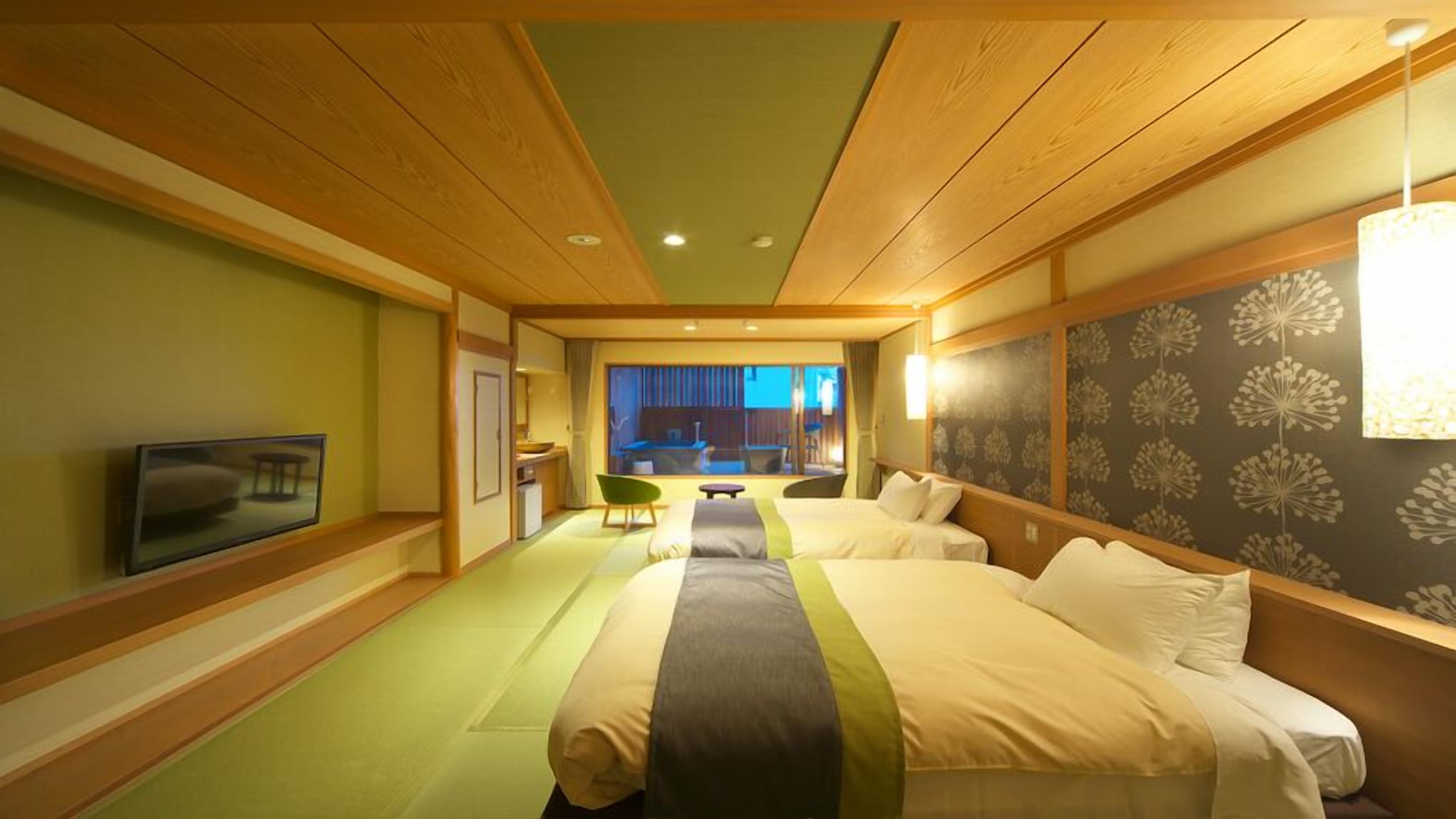 A hotel room with a bed, a television, a dining area, and a view of the cityscape.