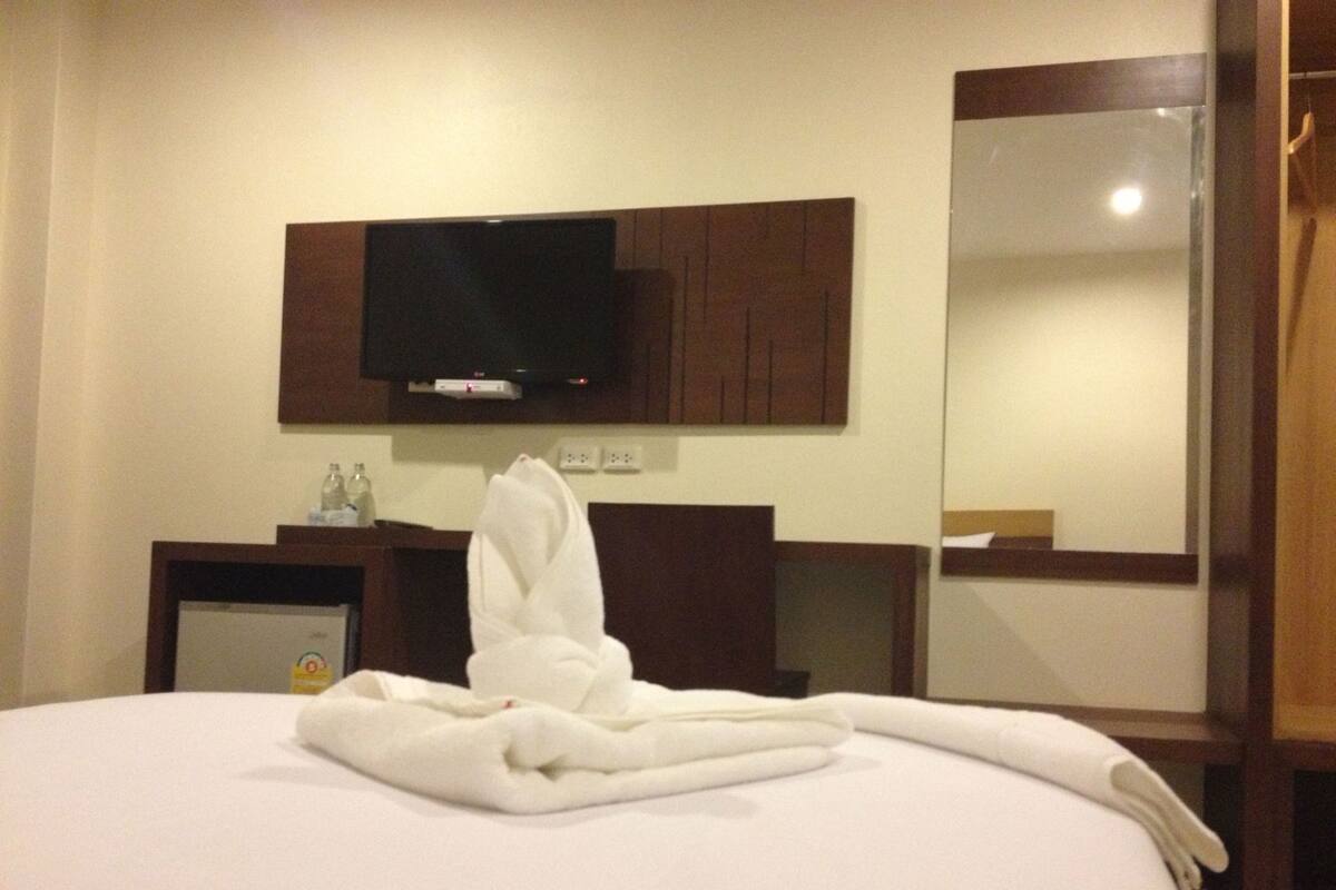 A hotel room with a bed, a television mounted on the wall, a mirror, and a small table with a bottle and a glass.