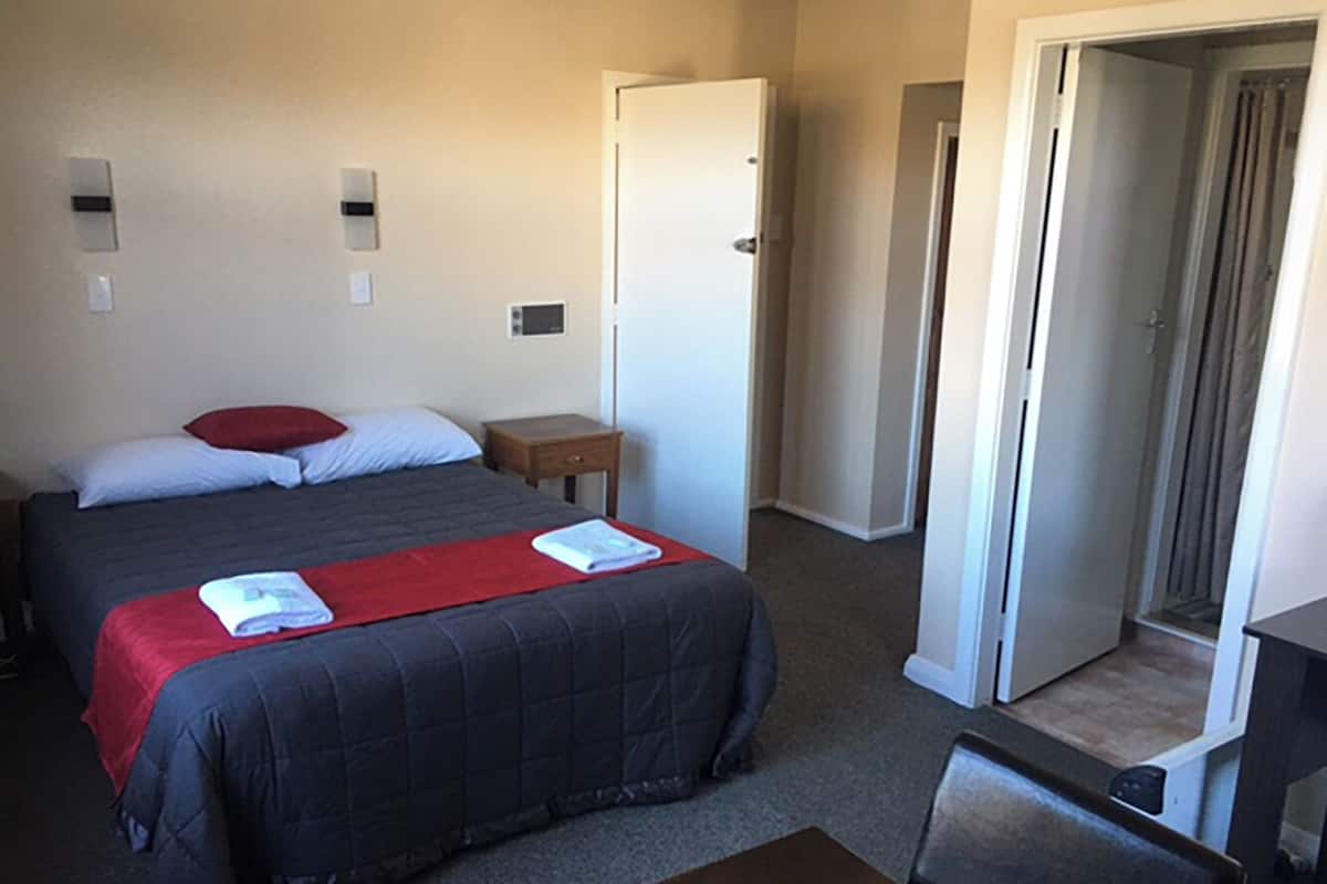 A hotel room with a bed, a small table, a chair, and a door leading to another room.