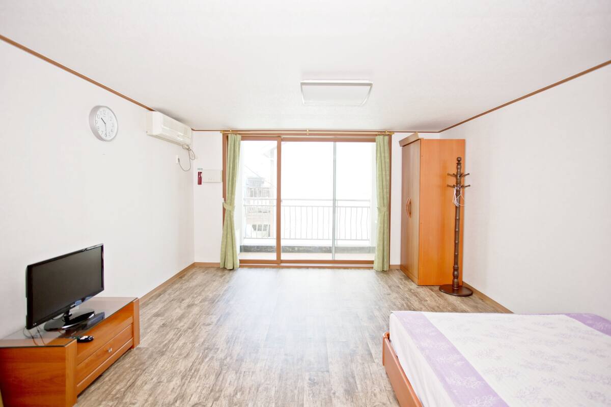 A room with a bed, a TV on a wooden dresser, a sliding glass door to a balcony, and a coat rack.