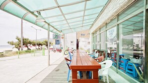 Ein Sitzbereich im Freien mit einem Holztisch und Stühlen, einem überdachten Gang und Blick auf das Meer.