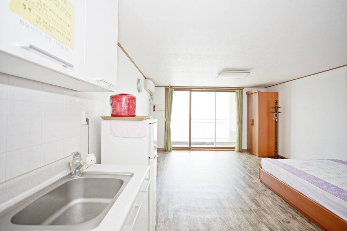 A small, bright room with a wooden floor, a bed, a sink, and a window with a view.