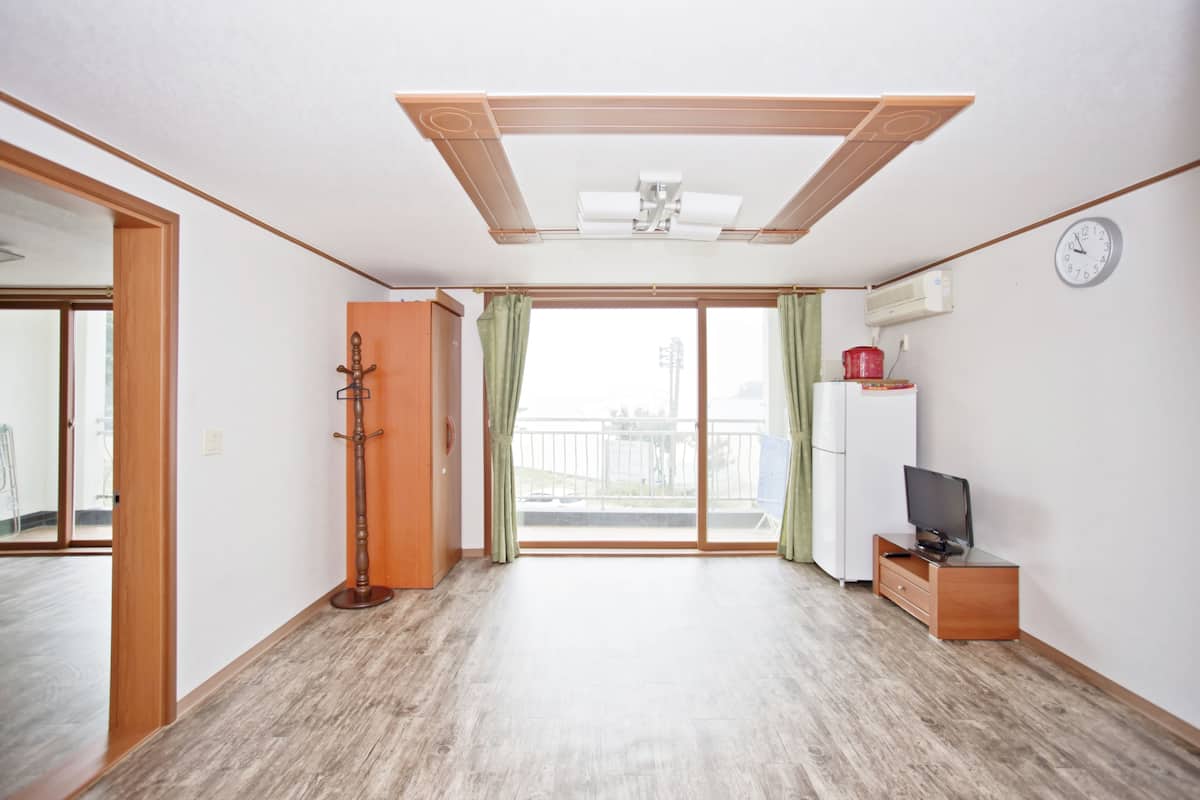 A spacious room with a large sliding glass door leading to a balcony, a ceiling fan, a small wooden cabinet, and a television on a stand.