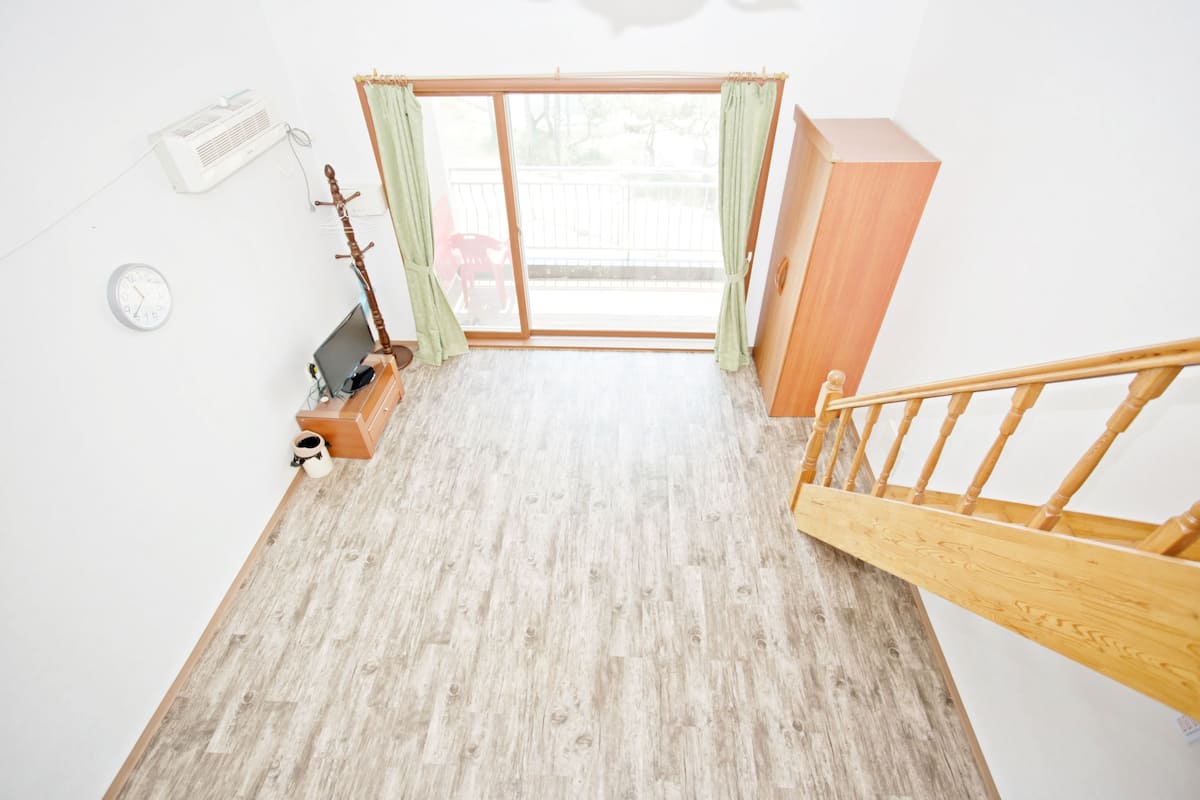 A room with a wooden floor, a wooden staircase, a sliding glass door, a small table with a chair, a clock, and an air conditioning unit.