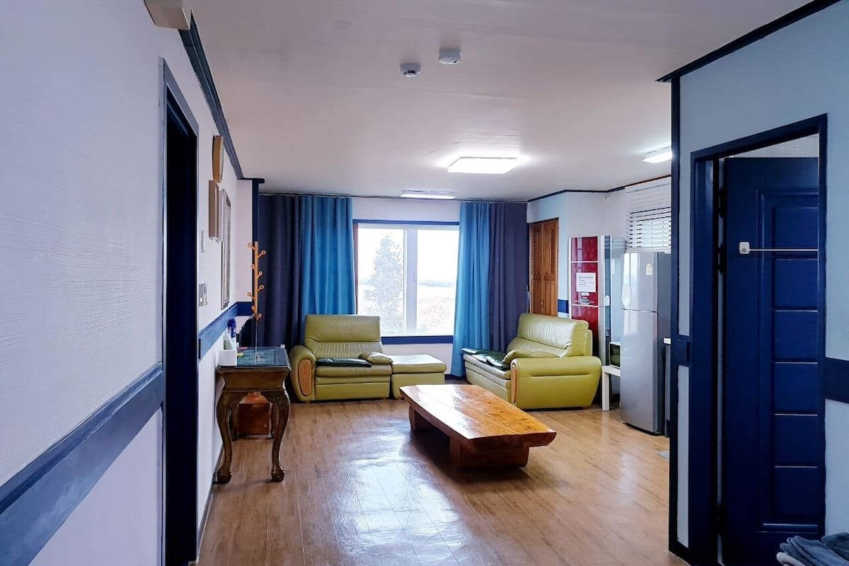 Ein Zimmer mit Holzboden, zwei gelbe Sofas, einem Holztisch, einem kleinen Tisch mit einer Lampe und einem Fenster mit blauen Vorhängen.