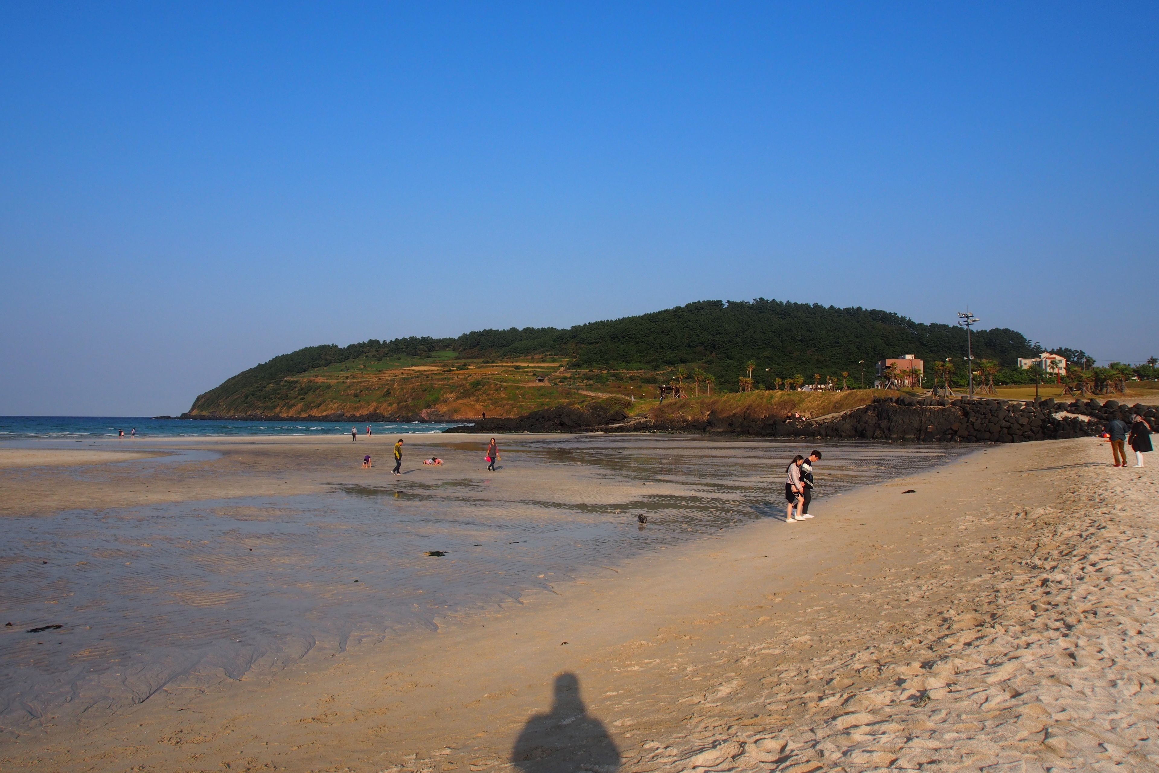 Una playa con gente caminando y parada, un promontorio rocoso y una colina al fondo.