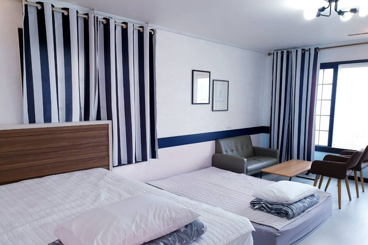 Ein Hotelzimmer mit zwei Betten, einem Schlafsofa, einem kleinen Tisch und einem Sessel. Vorhänge, ein Kopfteil und ein Fenster mit Blick sind vorhanden.
