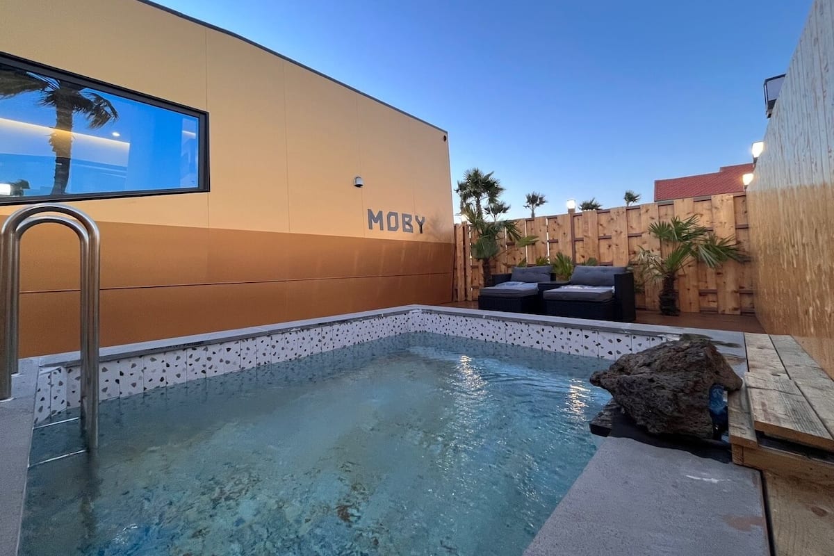 Ein Schwimmbecken mit Holzterrasse, eine Lounge mit Kissen und eine Wand mit dem Schriftzug 'MOBY'.