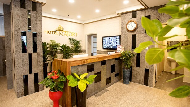 A hotel reception area with a reception desk, a clock, and a television displaying images. - Hotel Major 2 (Jeju City)