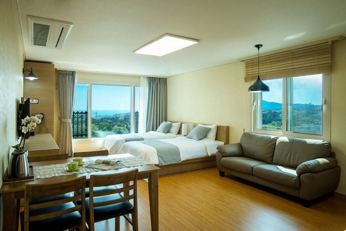Quarto de hotel com duas camas, um sofá, uma mesa de jantar com cadeiras e janelas amplas com vista.
