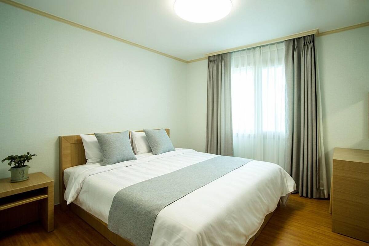 Quarto bem arrumado com cama, mesas de cabeceira, luminária e um vaso de planta, além de um espelho na parede.
