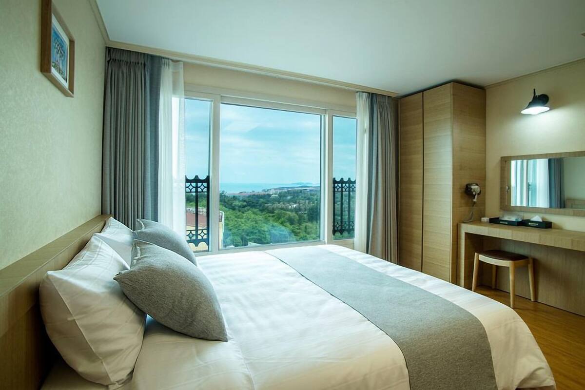 Quarto de hotel com uma cama grande, uma escrivaninha com cadeira, um espelho e vista para a paisagem urbana através de amplas janelas.