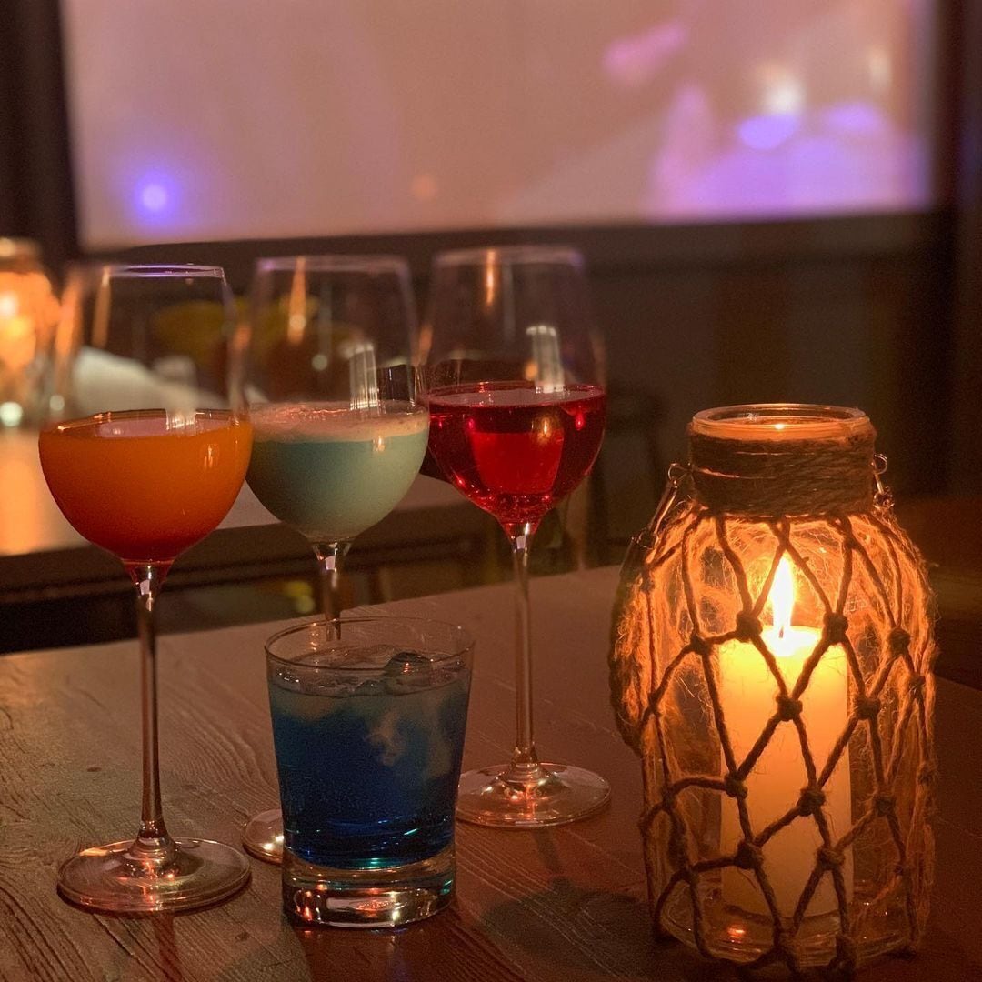 Una mesa con cuatro vasos de bebidas coloridas y una vela encendida dentro de un frasco de vidrio.