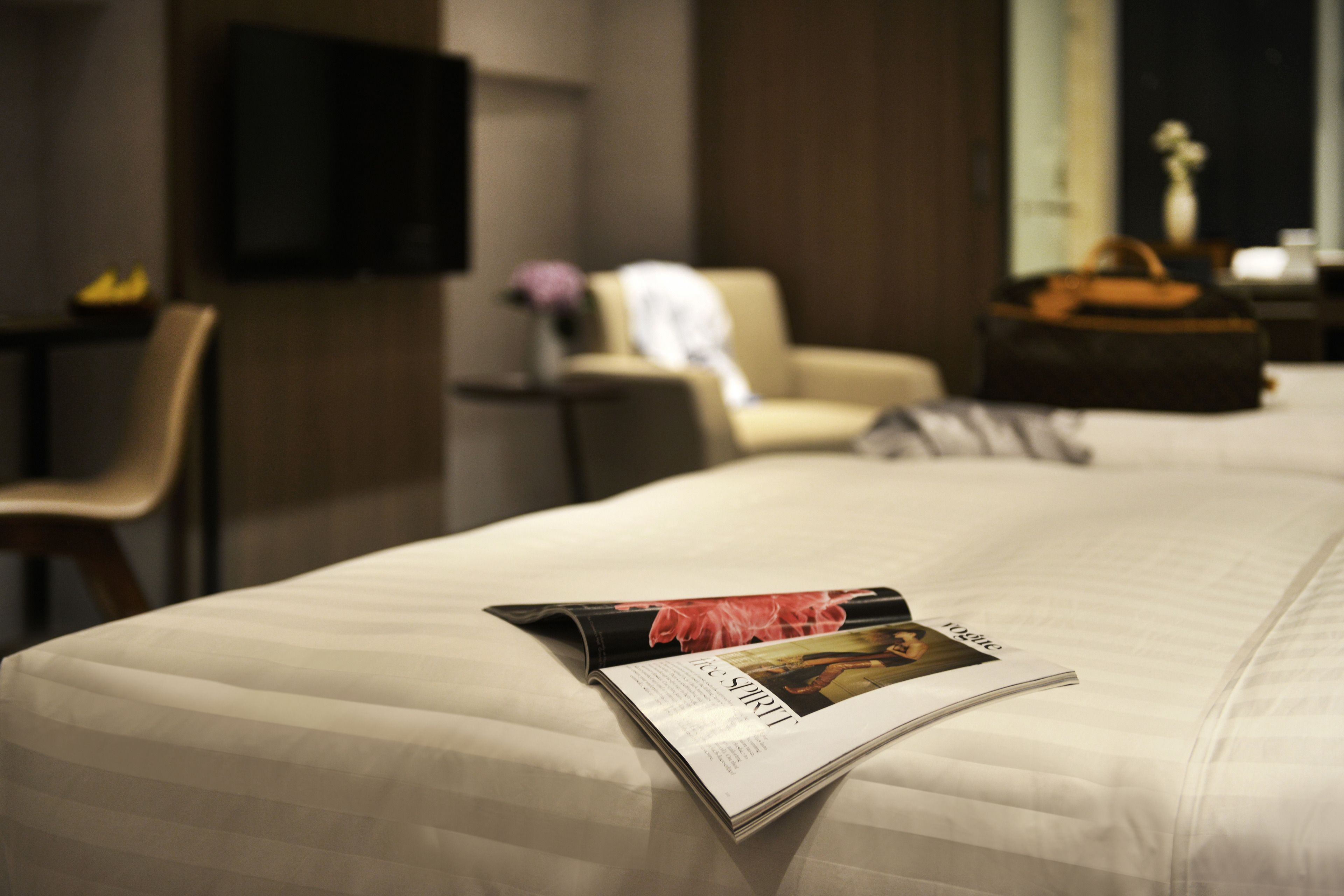 A neatly made bed with a magazine on it, a chair, a table with a vase, and a television on the wall.