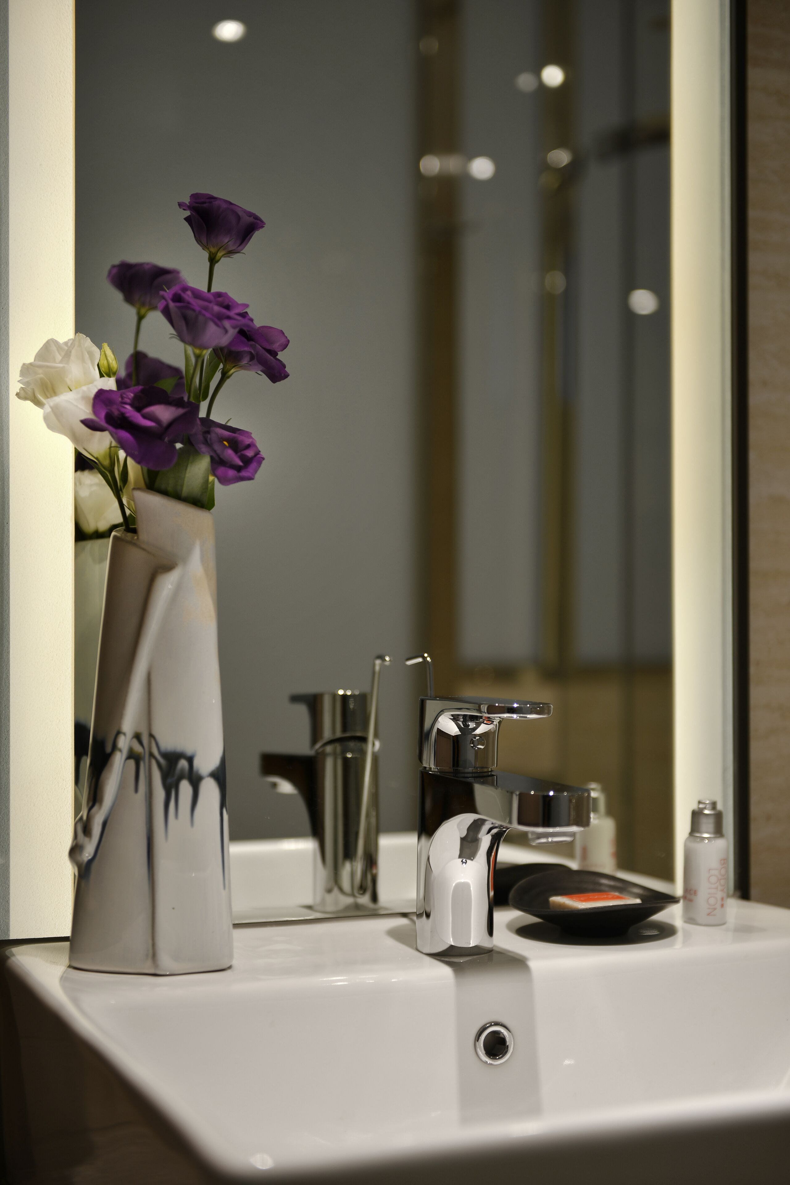 Un baño con un lavamanos blanco, un espejo y un florero con flores moradas y blancas.