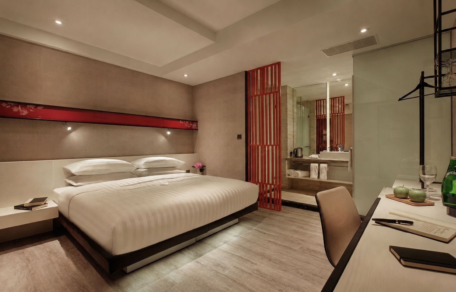 A modern hotel room with a large bed, bedside tables, a desk with a chair, a mirror, and a bathroom visible through a glass partition.