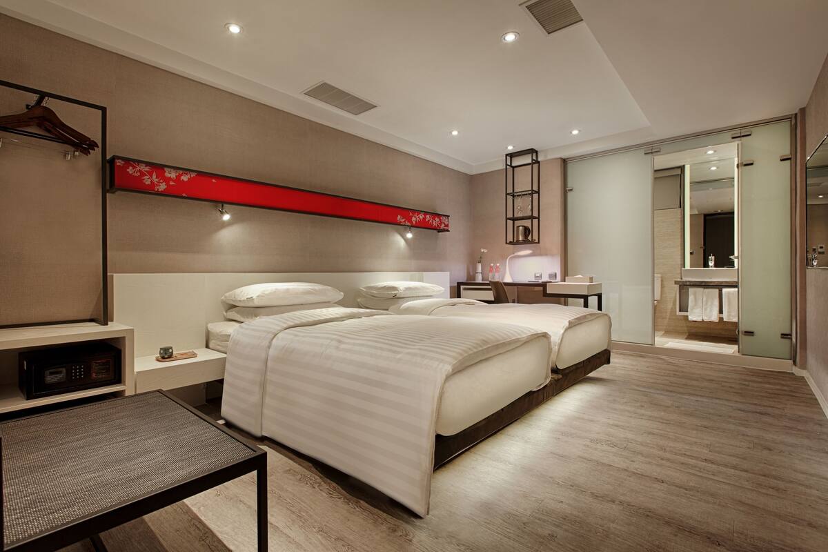 Ein modernes Hotelzimmer mit zwei Betten, einem Schreibtisch, einem Stuhl, einer roten Wandverkleidung und einem Badezimmer mit Spiegel und Handtuchhalter.