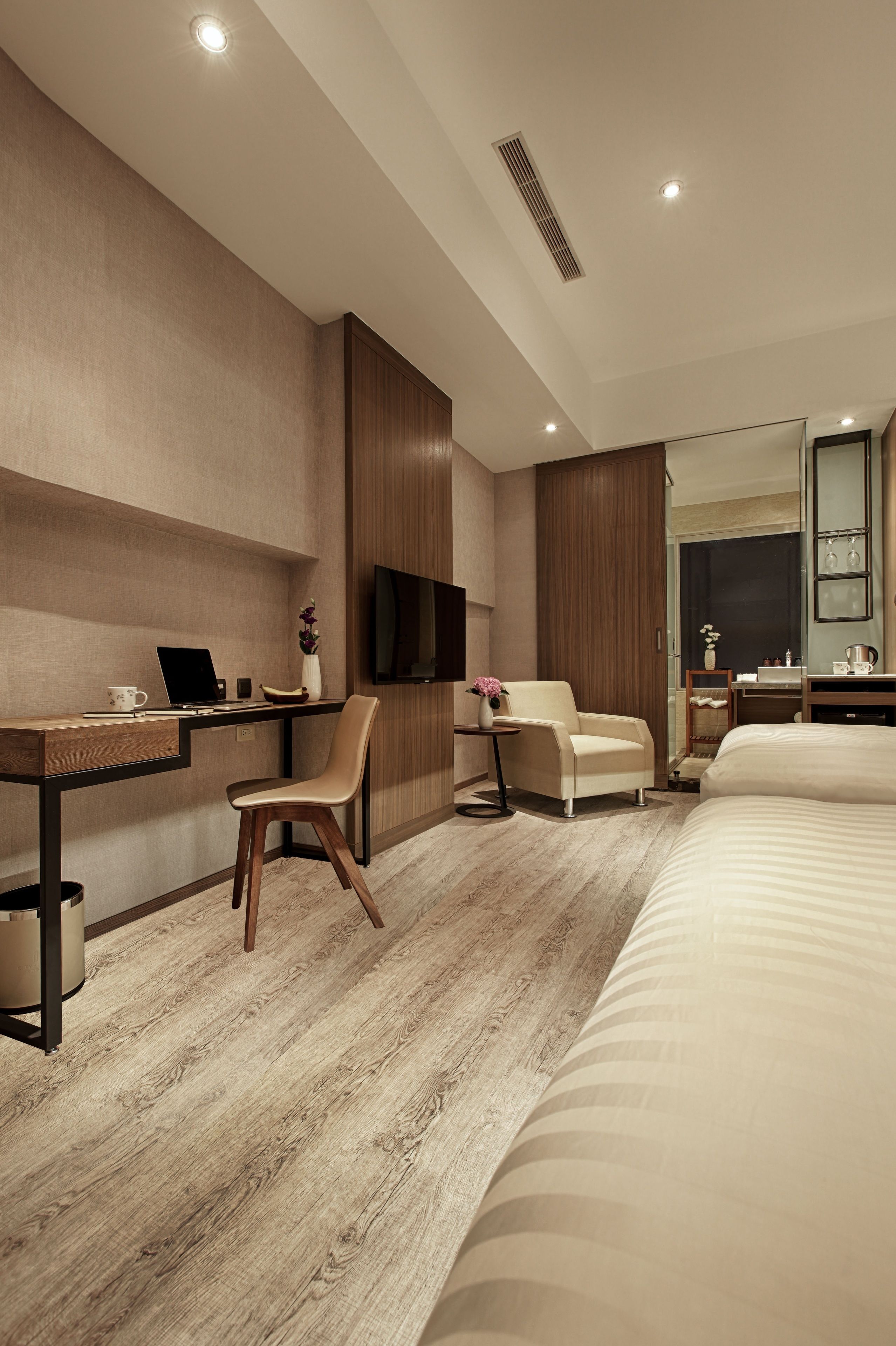 Una habitación de hotel moderna con un escritorio de madera, una silla cómoda, una cama con un espejo grande y un televisor montado en la pared.
