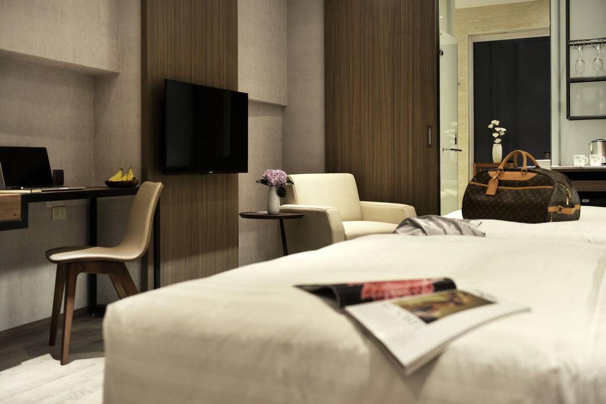 Ein Hotelzimmer mit einem Bett, einem Schreibtisch mit einem Laptop, einem Sessel, einem Fernseher und einem kleinen Tisch mit einer Vase und einer Tasche.