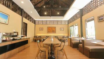 A spacious dining area with a long wooden table surrounded by chairs, a high ceiling with exposed beams, and large windows allowing natural light.