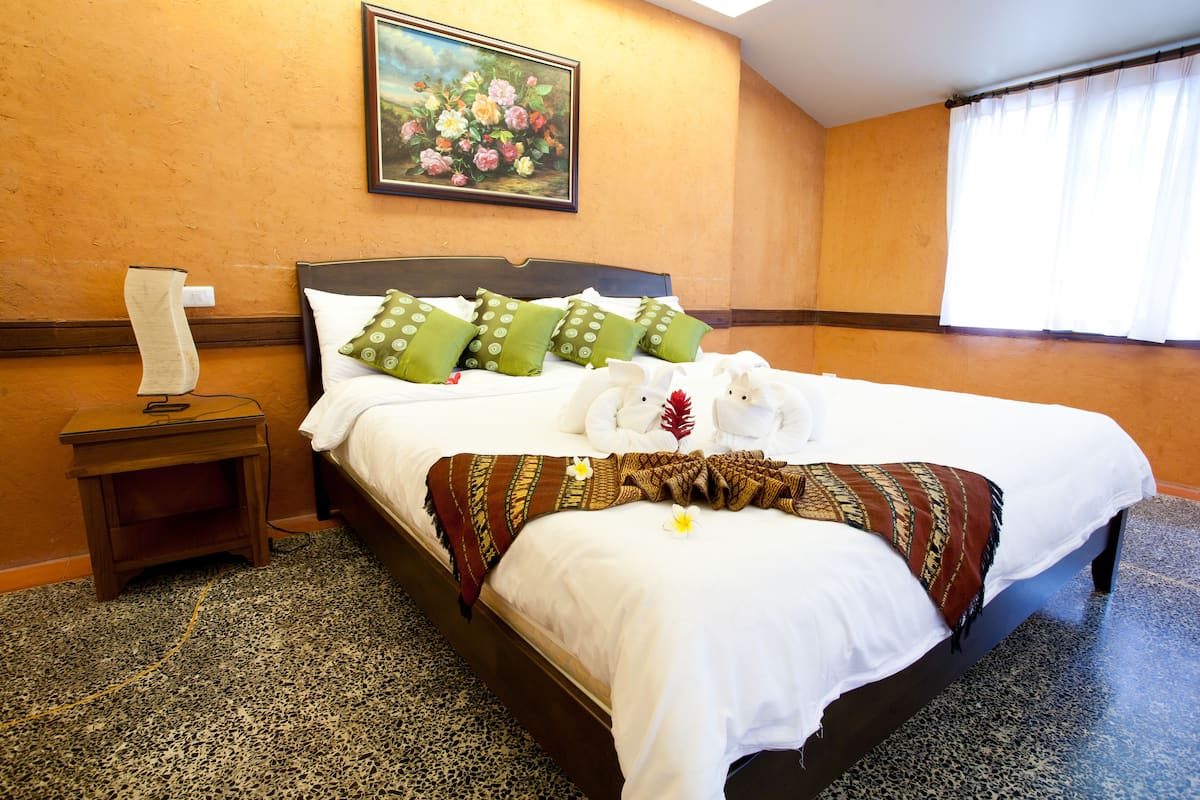 A neatly made bed with white linens and green pillows, a painting of flowers on the wall, and a small wooden nightstand with a lamp.