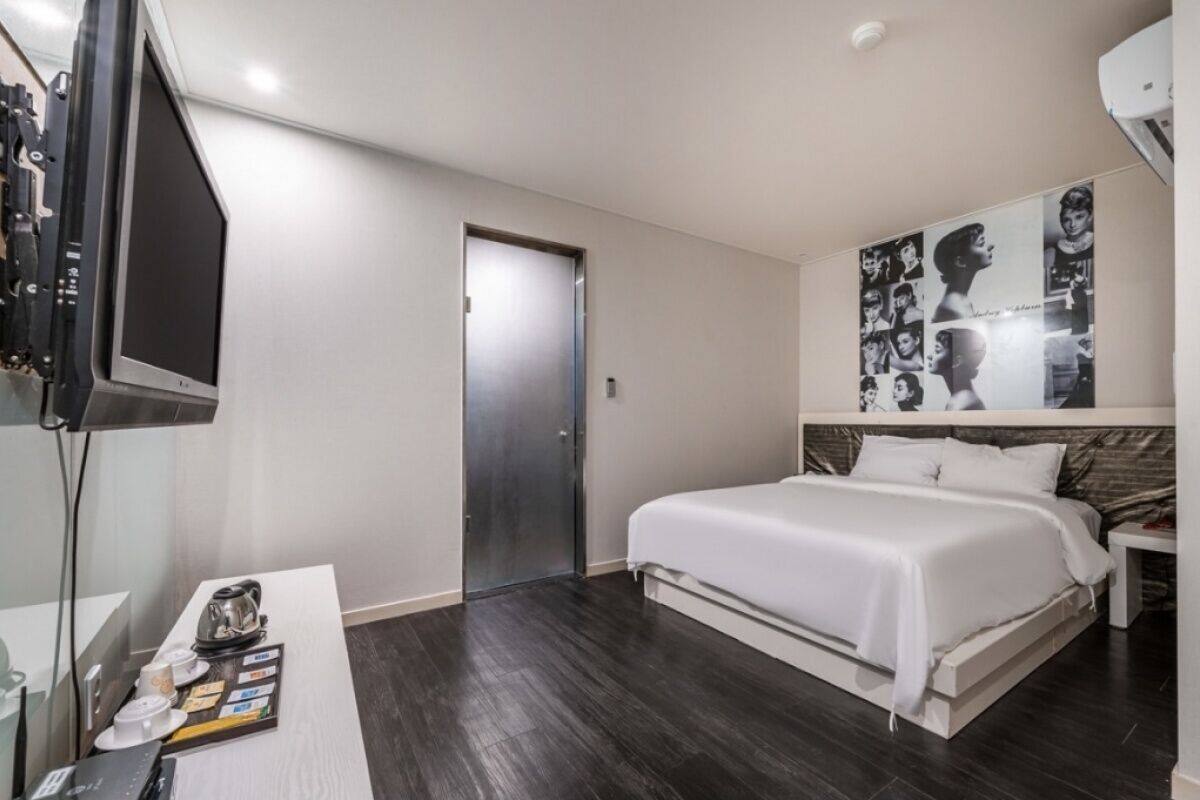 A modern hotel room with a bed, a desk with a kettle, a television, and a wall with a poster.