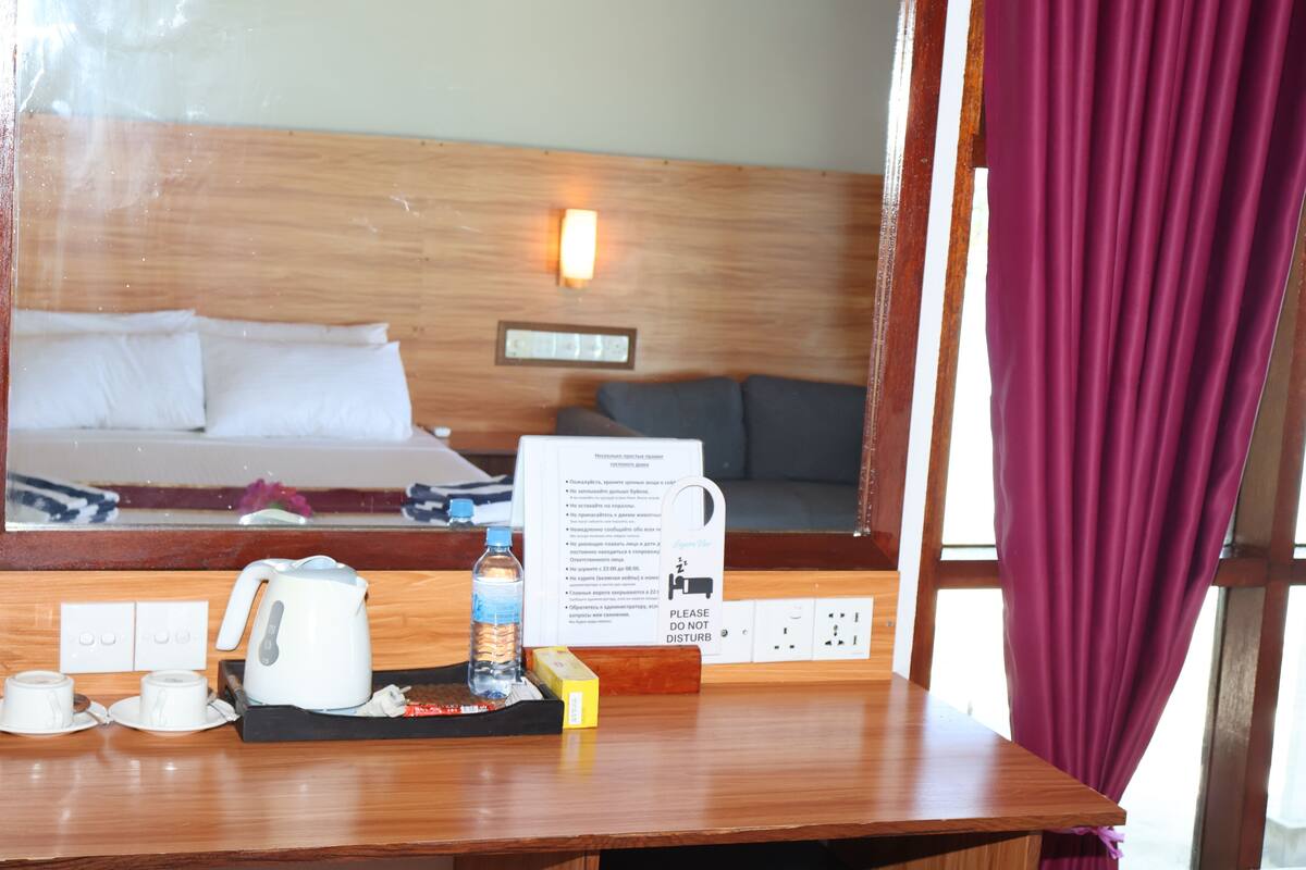 Quarto de hotel com cabeceira de madeira, mesa de cabeceira com chaleira, chávenas e garrafa de água. Um aviso na parede fornece informações sobre o quarto.