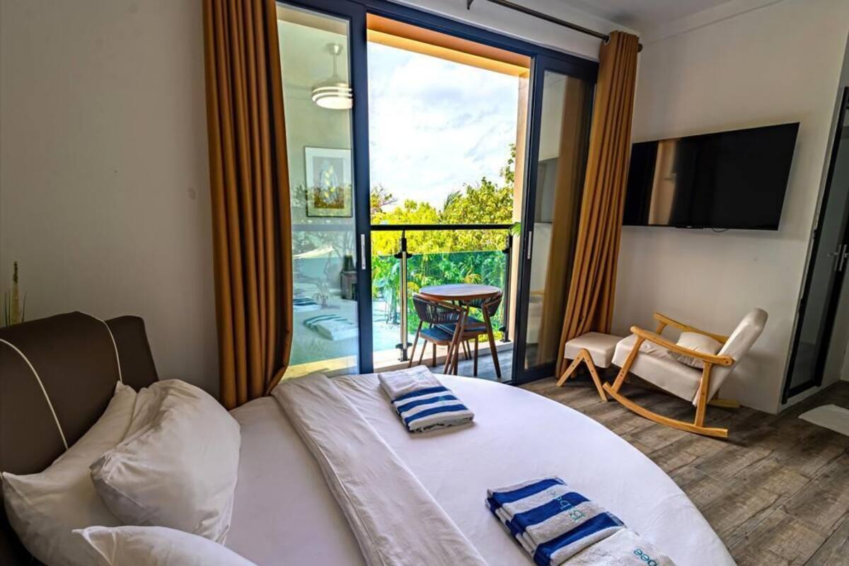 A hotel room with a bed, a chair, a table, and a balcony with a view of greenery.