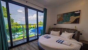 A modern hotel room with a large bed, a painting on the wall, and a balcony with a view of greenery and a blue-roofed building. - Bibee Maldives (Dhiffushi)