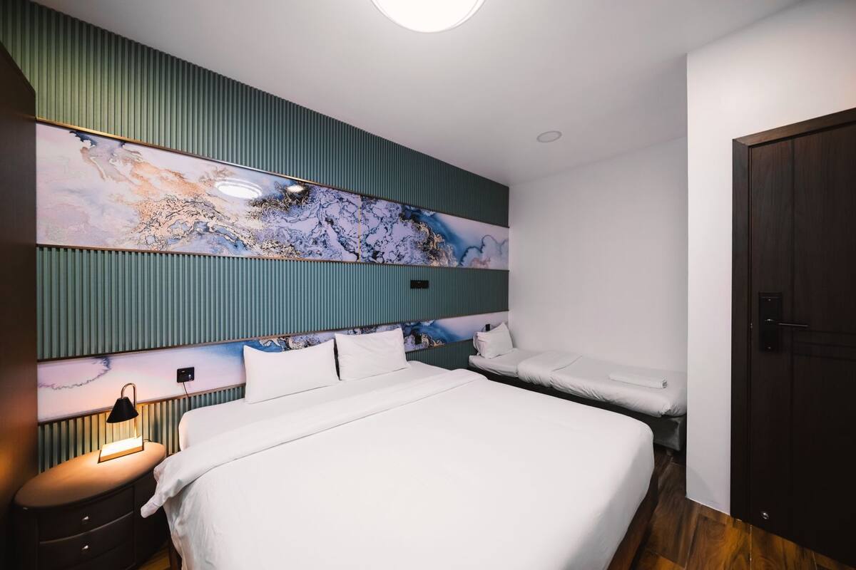 A modern hotel room with a large bed, a bedside table with a lamp, a headboard with a decorative panel, and a door.