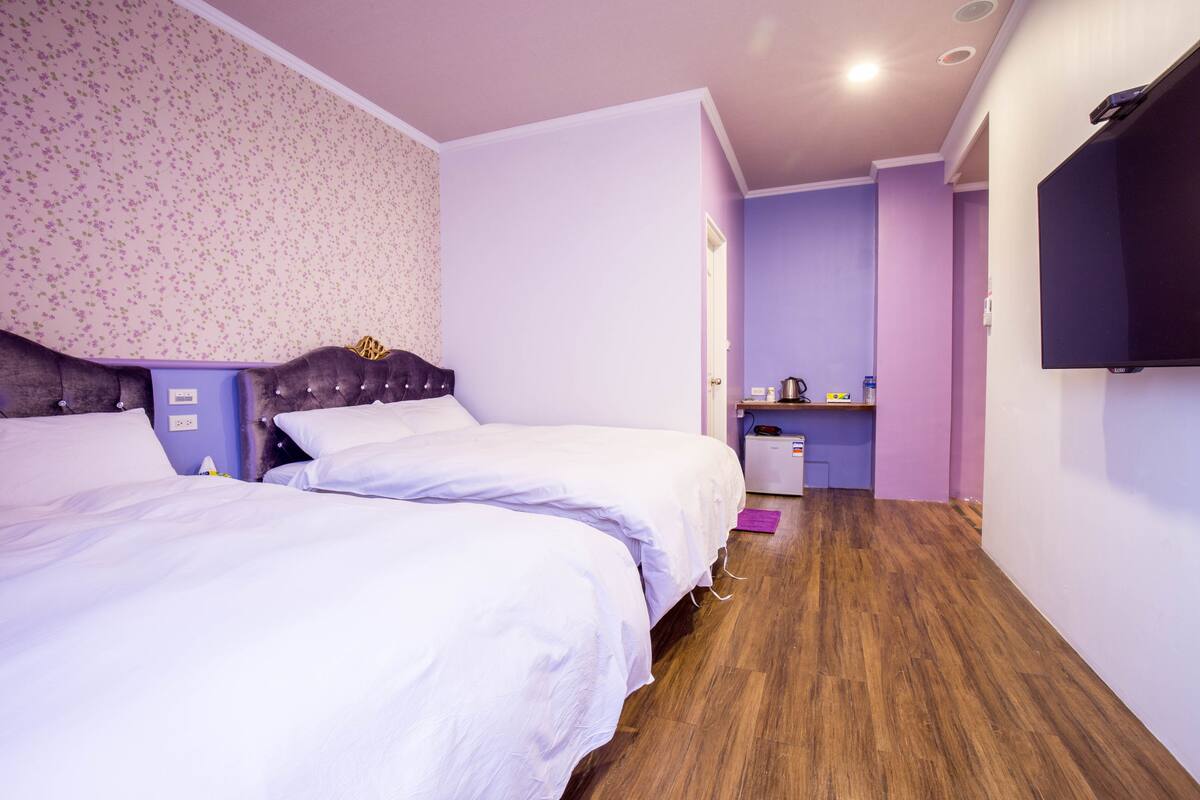 Habitación de hotel con dos camas, suelo de madera, una pequeña cocina americana y un televisor montado en la pared.