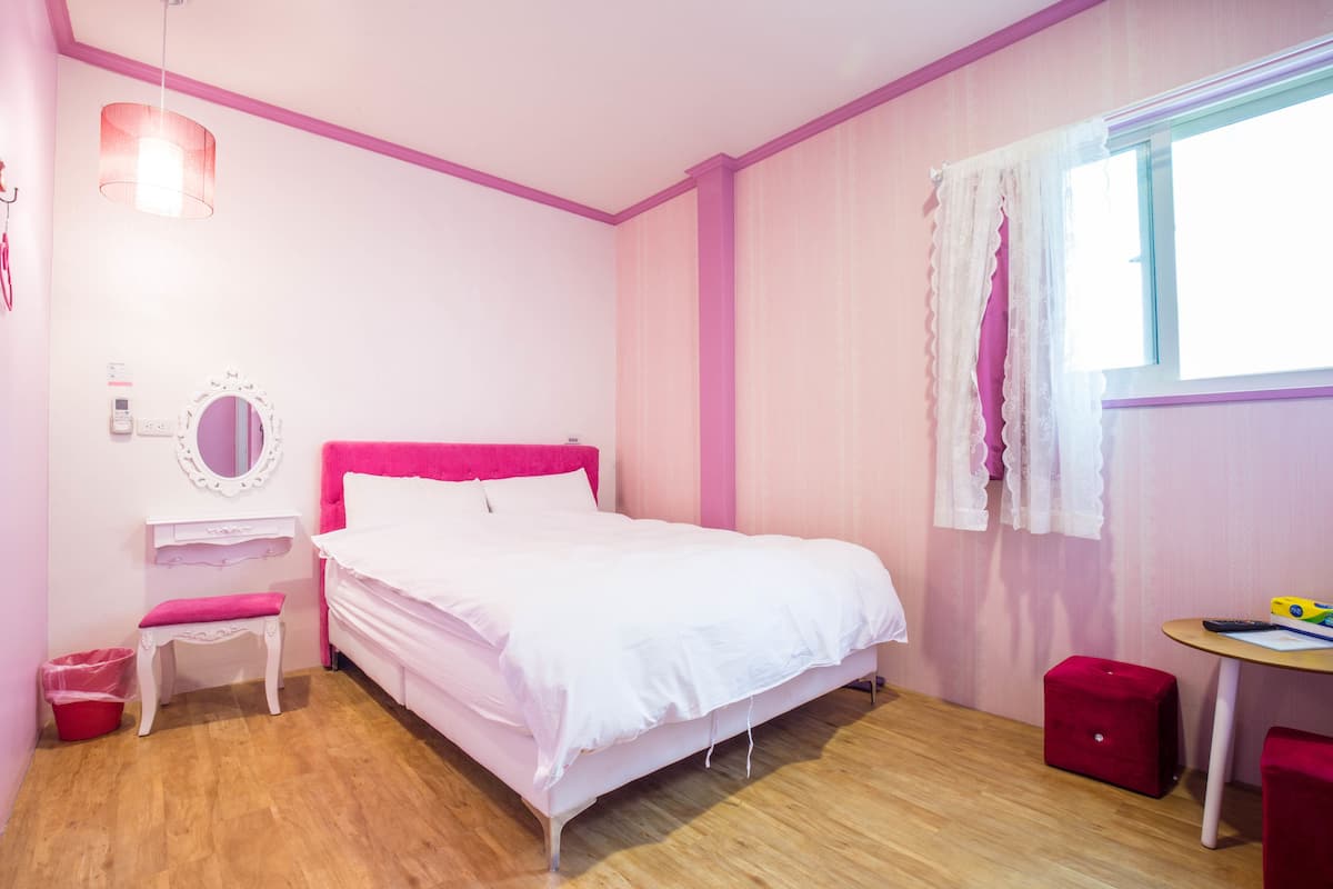 Un dormitorio con temática rosa, que incluye una cama, un tocador, un espejo, un taburete pequeño y una ventana con cortinas.