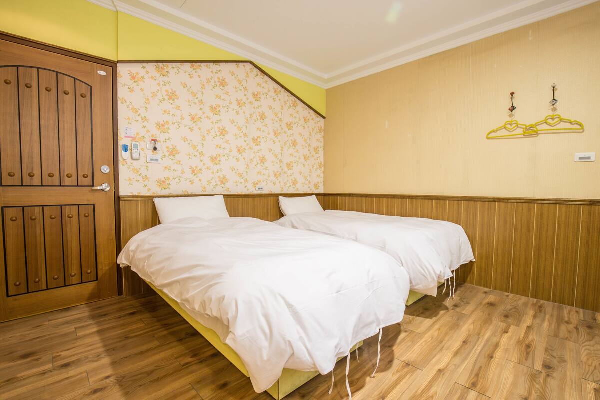 Una habitación con dos camas, paneles de madera, un área con papel tapiz floral y una puerta de madera.