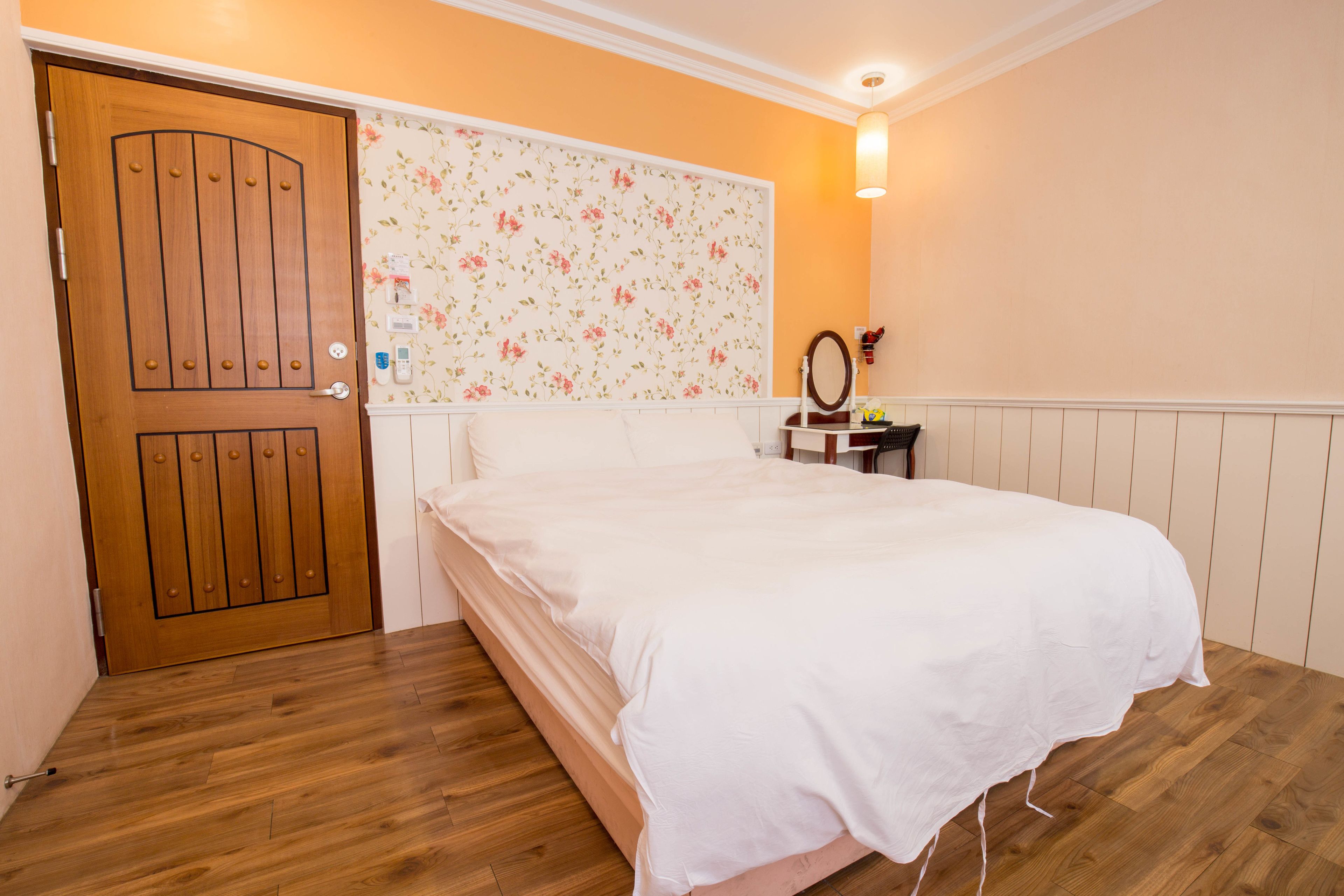 Une chambre avec une porte en bois, un lit avec une literie blanche, une tête de lit à motifs floraux, une table de chevet avec un miroir et un luminaire fixé au mur.