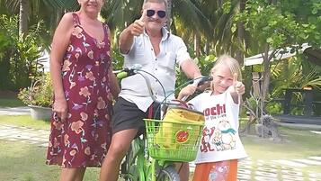 A family on a green bicycle with a basket, standing in a garden with palm trees.